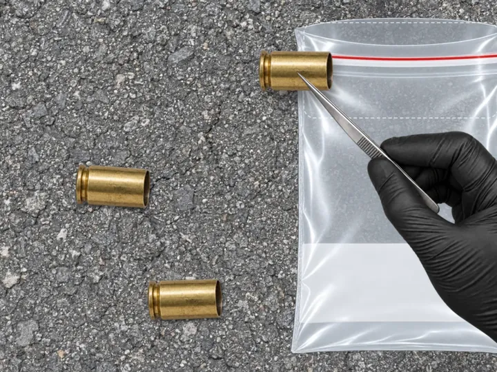 Brass shell casings on asphalt, a gloved hand holding one in tweezers over an evidence bag
