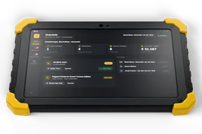 A rugged black tablet with yellow corner bumpers lying screen up, its screen showing the workshop app overview with open calls, work orders, today's invoices and the colleagues on duty